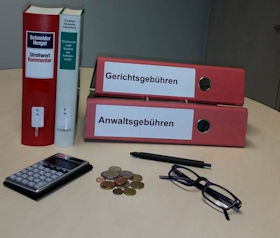 Das Bild zeigt zwei stehende Kommentare Streitwert und zwei liegende Ordnern Gerichtsgebühren, Anwaltsgebühren im Hintergrund, vorne im Bild links ein Taschenrechner, in der Mitte einige Geldmünzen sowie rechts eine Brille und Kugelschreiber.